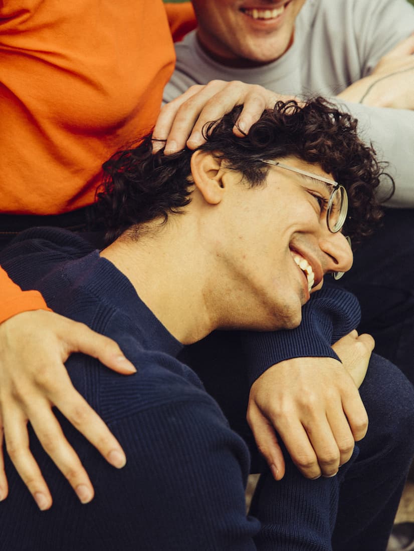 A smiling person with curly hair and glasses leans on another's knee, surrounded by hands. People are wearing casual clothes in warm colors.